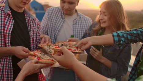 A-company-of-six-young-russian-people-in-plaid-shirts-parses-pieces-of-hot-pizza-with-red-paper-and-eats-together.-This-is-a-rooftop-party-with-a-beer.